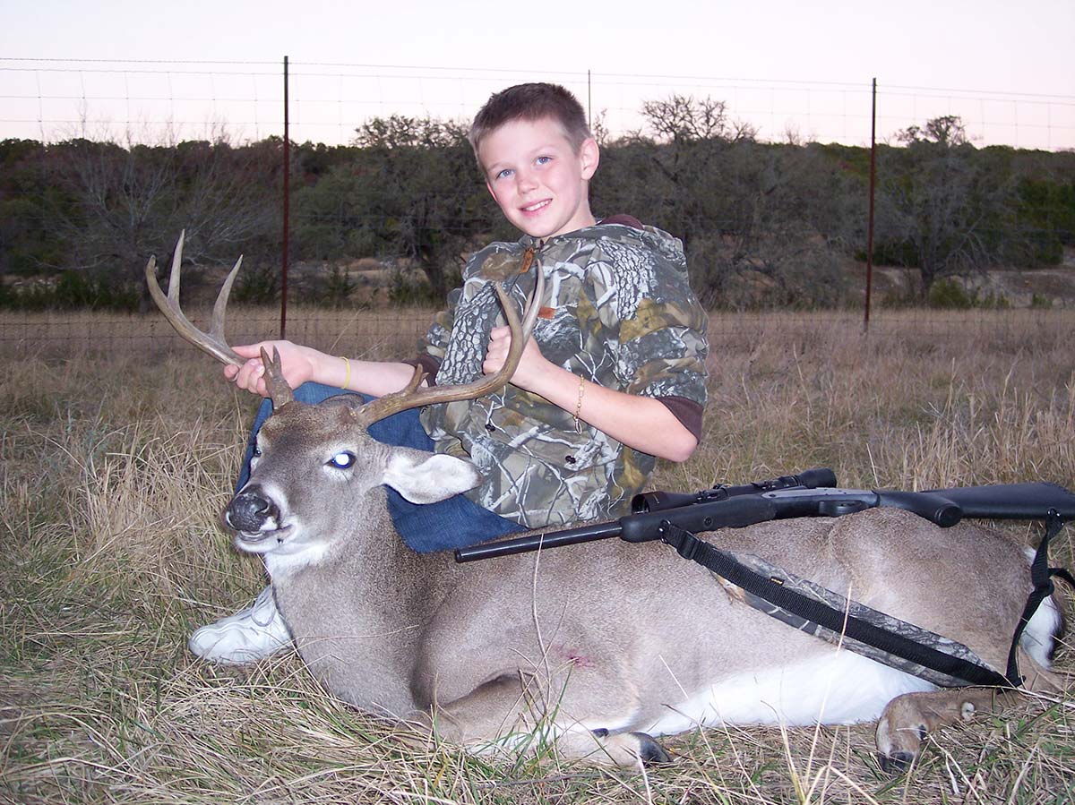 Ashton in full camo kneeling behind a whitetail buck in a dusk field, rifle across the deer