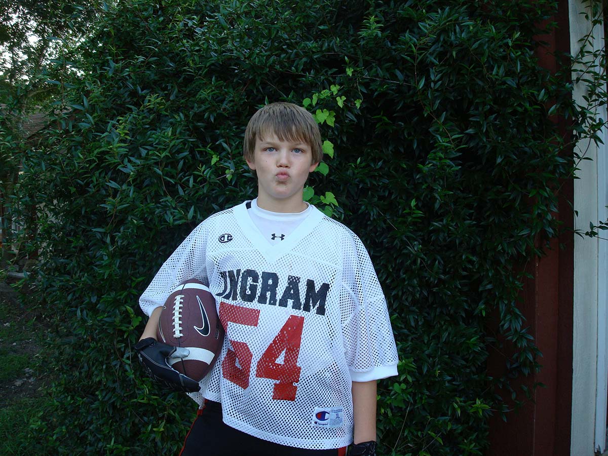 Ashton standing against the ivy in a white Ingram number 54 football jersey, football tucked under one arm