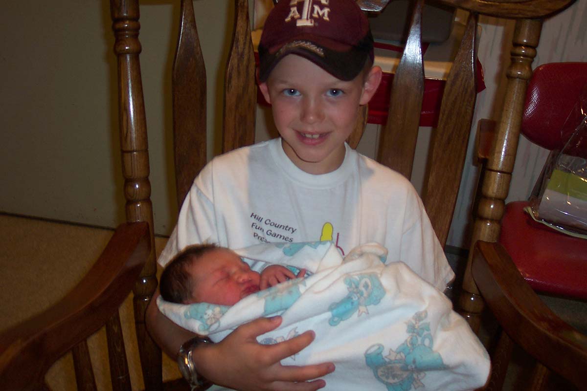 Ashton in a Texas A&M cap at the kitchen table, very carefully holding his newborn baby brother Jayden