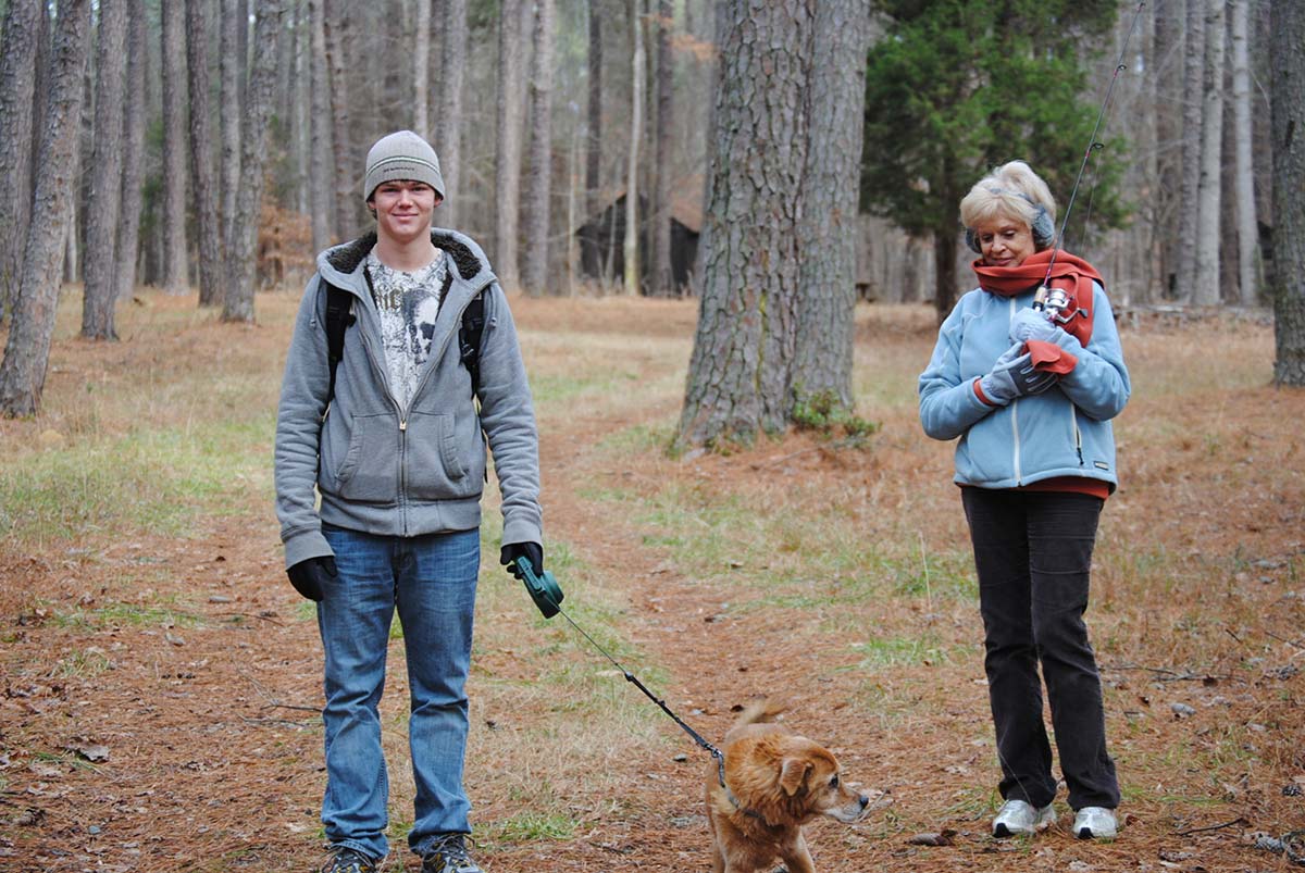Ashton in a gray hoodie and beanie walking his dog on a pine-woods trail, his Grandmother Love beside him cradling a fishing rod