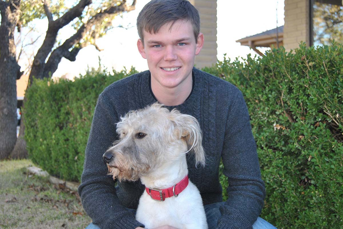 Ashton in a gray cable-knit sweater kneeling in front of a green hedge, holding his white dog and smiling into the camera