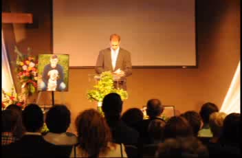 Speaker at the podium beside Ashton's framed portrait and a spray of red and white flowers, congregation seated
