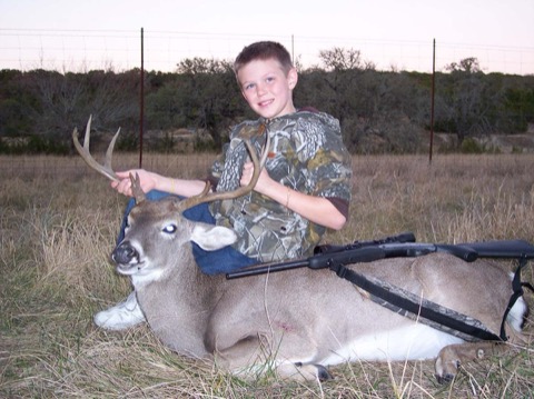 Ashton in full camo kneeling behind a whitetail buck in a dusk field, rifle across the deer