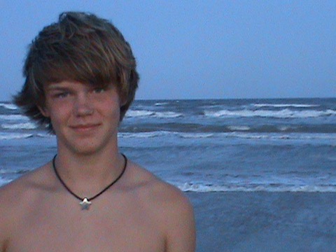 Ashton at the beach at dusk, star pendant on a black cord, ocean breaking behind him