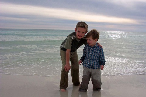 Young Ashton steadying his little brother Jayden ankle-deep at the shoreline under a huge luminous silver-and-blue sky