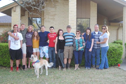 Twelve friends and family shoulder-to-shoulder on the front lawn, Ashton mid-line in the striped tee, his white dog wearing a stars-and-stripes bandana